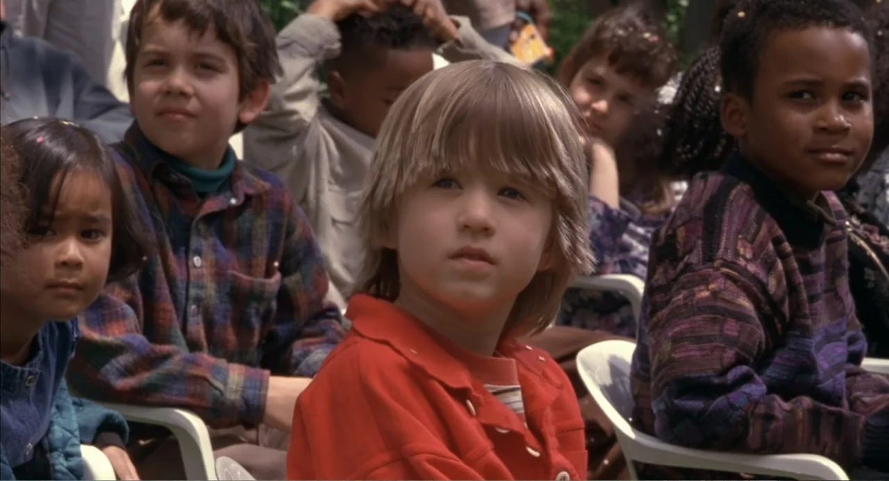 Bogus (1996) movie still: medium — A group of diverse children sit in plastic white chairs during an outdoor gathering. The central blonde boy faces…