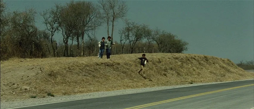 The Headless Woman (2008) movie still: wide — A dry, grassy roadside embankment borders a strip of paved asphalt road. Three figures run down the…