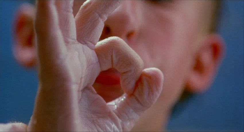 Gummo (1997) movie still: closeup — A close-up view shows a hand forming an okay sign against a blue wall. The person's face is visible in the…