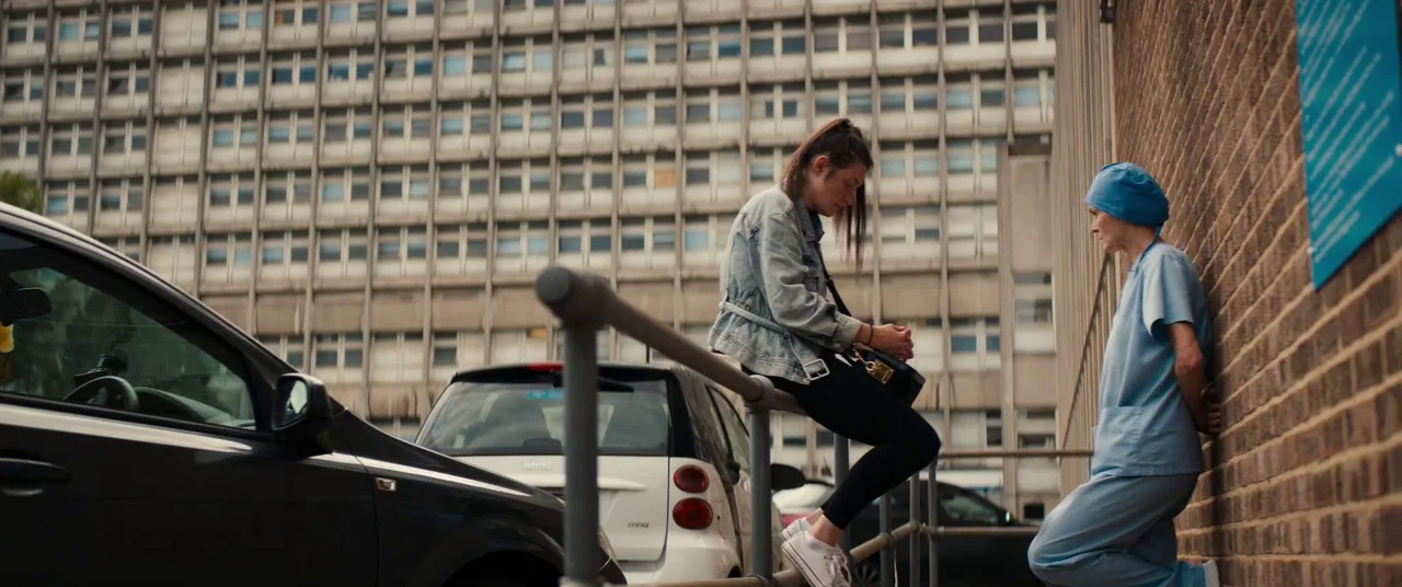 Silver Haze (2023) movie still: fullbody — A hospital parking lot sits beneath a large, industrial-style concrete medical building facade. A young…