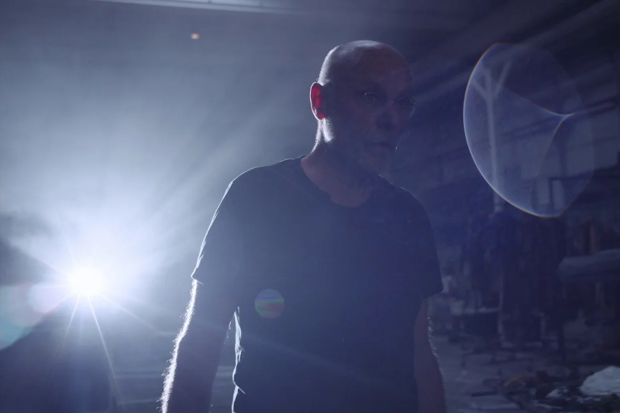 Anselm (2023) movie still: medium — A dark industrial space filled with hazy air and indistinct scattered equipment. A bald man stands in the…