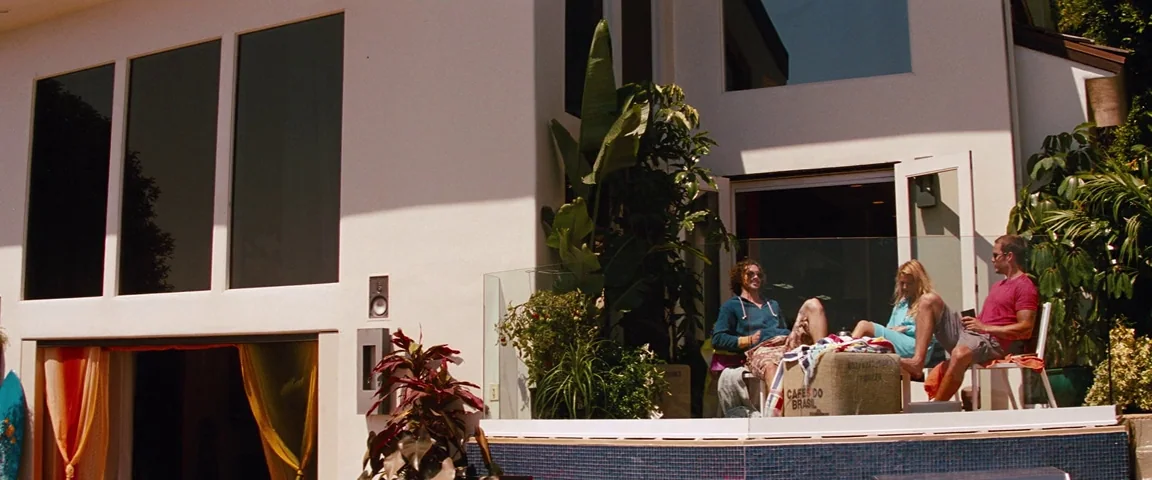 Savages (2012) movie still: wide — A modern white house features large windows and a small elevated patio terrace. Three people sit on chairs on the…