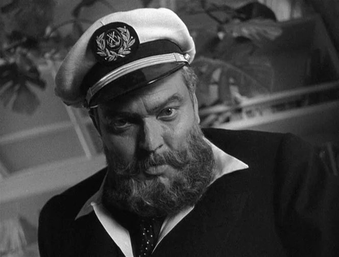 Mr. Arkadin (1955) movie still: closeup — A man wearing a nautical officer hat sits in a dim indoor space. The figure looks directly into the lens…