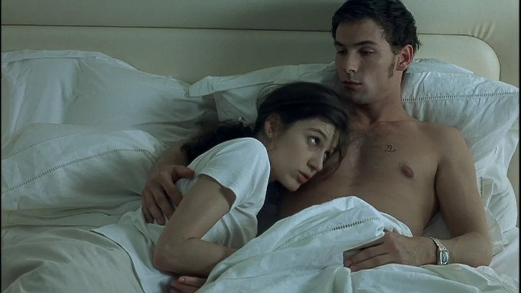 Romance (1999) movie still: medium — A pair of individuals lie together on a white unmade bed with plush pillows. The woman rests her head against…