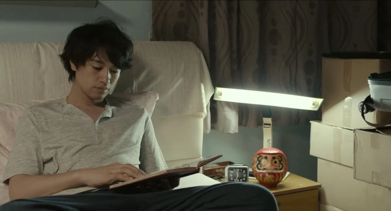 Ramen Shop (2018) movie still: medium — A bedroom corner features a headboard, a bedside table with a lamp, and cardboard boxes. A man wearing a…