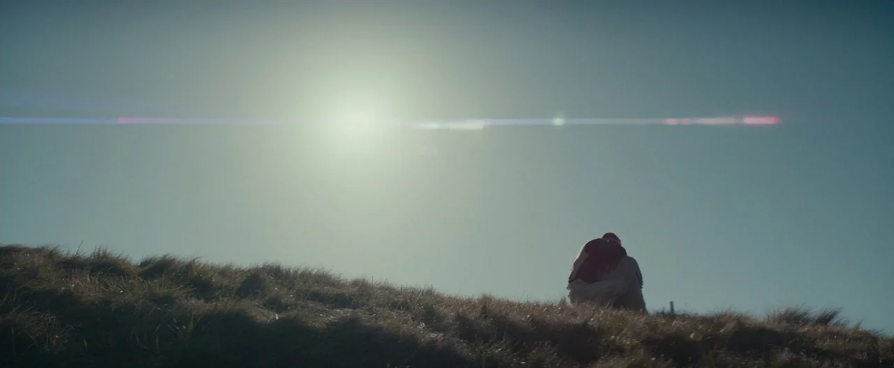 The Light Between Oceans (2016) movie still: wide — A grassy hill crests against a bright sun in a clear sky. Two figures sit closely together on…