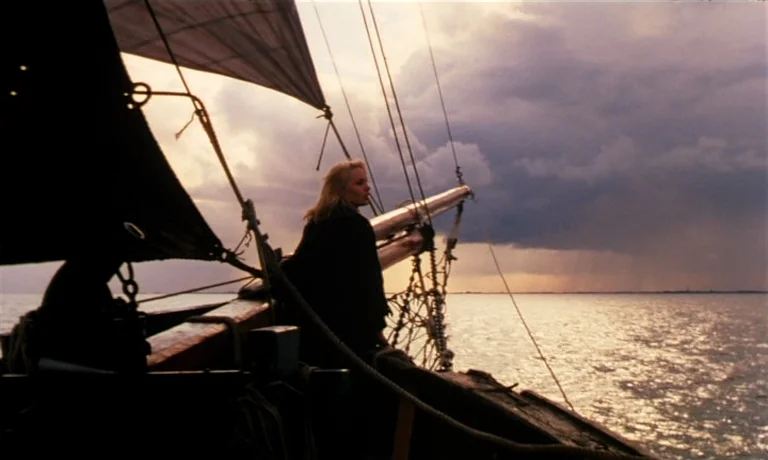 Katie Tippel (1975) movie still: medium — A young woman stands on the wooden deck of a sailboat at sunset. A dark, stormy cloud bank looms over the…