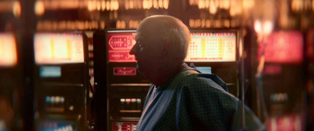 Elvis (2022) movie still: medium — An elderly man stands in profile inside a dimly lit area with slot machines. The background is filled with…