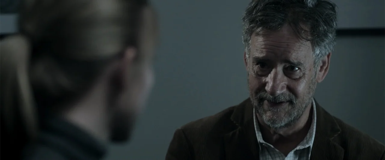 Nocturne (2020) movie still: medium — A dimly lit room provides a workspace with simple wall surfaces. An older man with a graying beard turns his…