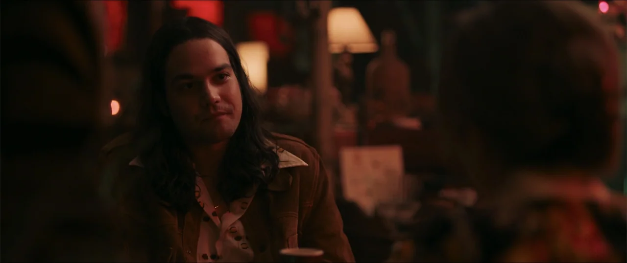 Woman of the Hour (2024) movie still: medium — A dimly lit indoor space with blurred furniture and a warm lamp in the background. A young man with…