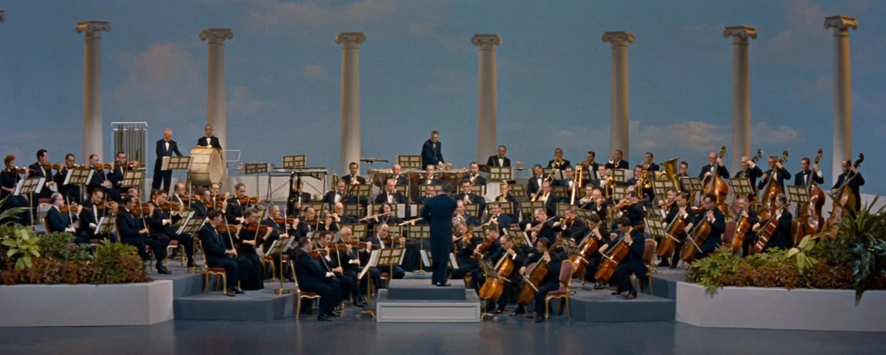 How to Marry a Millionaire movie still: wide — A large symphony orchestra performs on a stage decorated with white classical columns. The conductor…