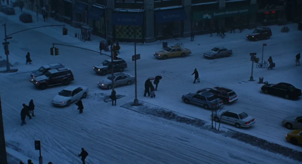 Mimic (1997) movie still: wide — A snowy urban intersection at blue hour features multiple vehicles and pedestrians moving slowly. Tall buildings…