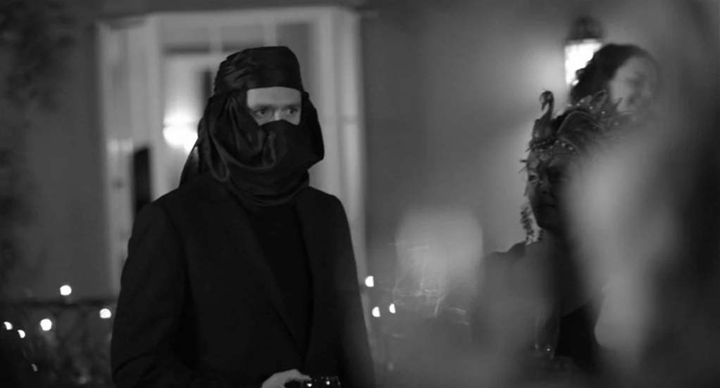 Much Ado About Nothing (2012) movie still: medium — A dimly lit ballroom with figures in masquerade attire gathered during a social event. A man…