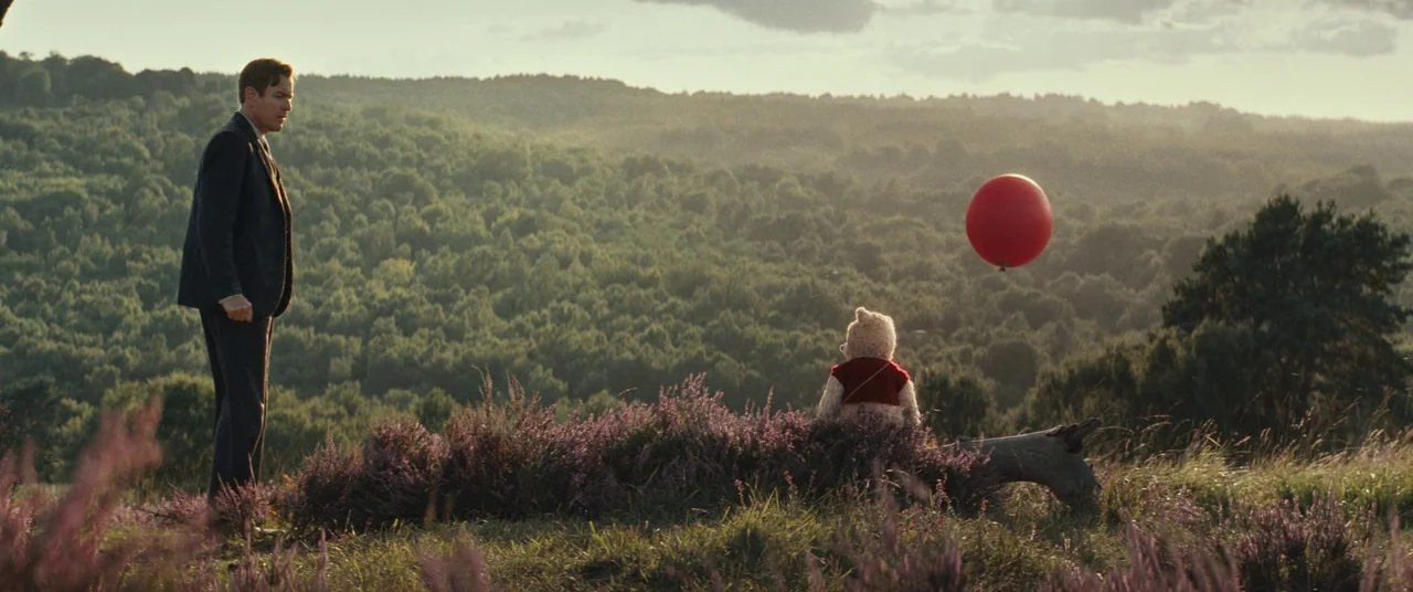 Christopher Robin (2018) movie still: wide — A man stands on a rolling green hillside covered in patches of purple heather. He faces a plush bear…