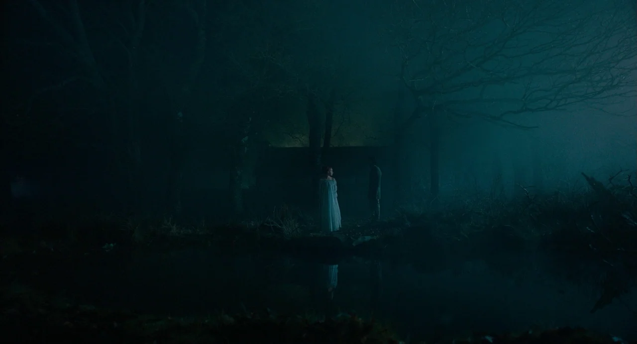 The Green Knight (2021) movie still: wide — Two figures stand on the edge of a dark, murky pond at night. The surrounding landscape is enveloped in…
