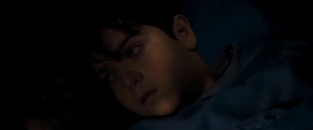 Baskin (2015) movie still: closeup — A dimly lit room at night provides a dark background for the subject. A young boy lies on his side, his face…