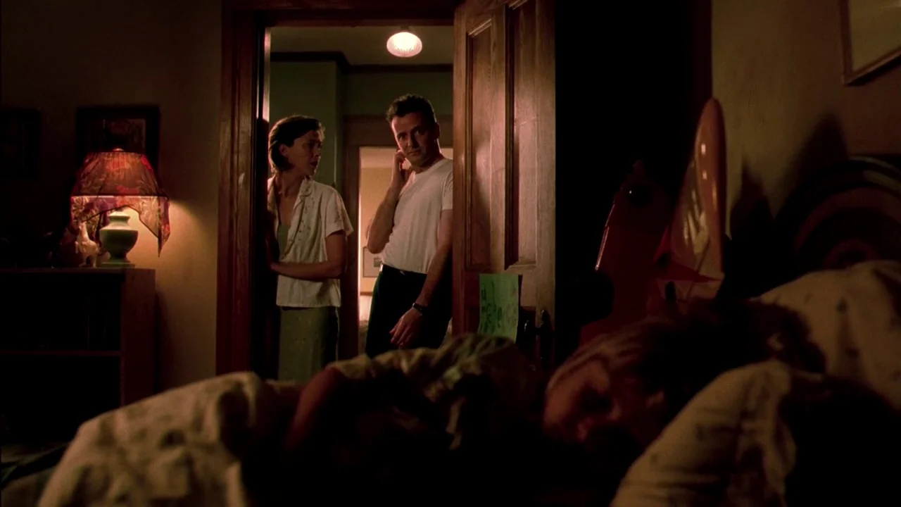 In Dreams (1999) movie still: medium — A dimly lit room at night with a sleeping figure in the foreground. A man and woman stand together in a…