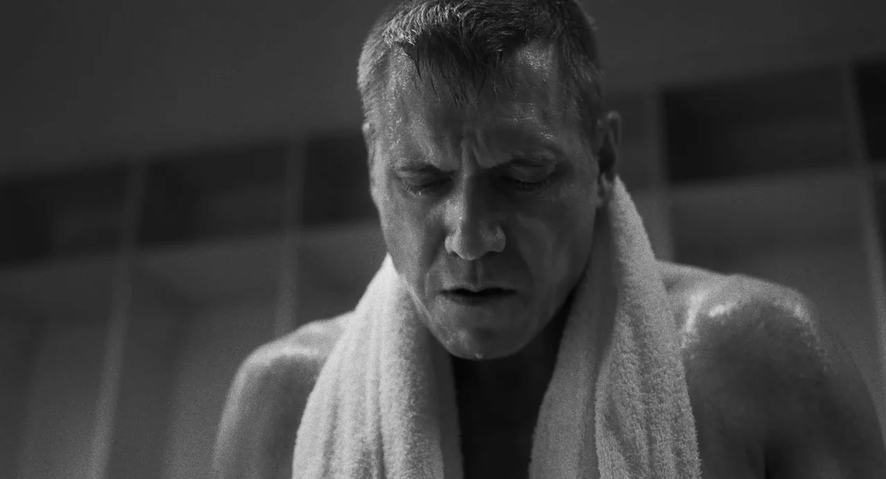 The Iron Claw (2023) movie still: closeup — A locker room with white lockers visible in the blurred background. A shirtless man with a white towel…