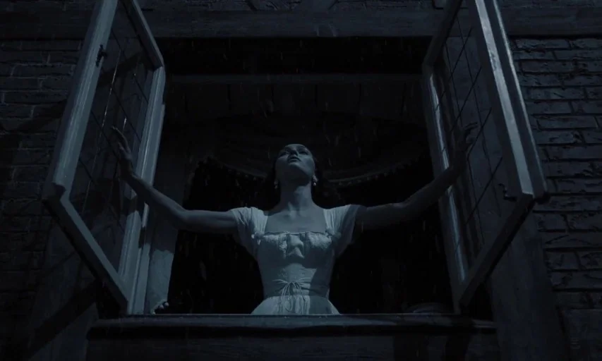 Nosferatu (2024) movie still: medium — A woman stands in a rectangular window opening at night during a rain shower. She reaches outward with both…