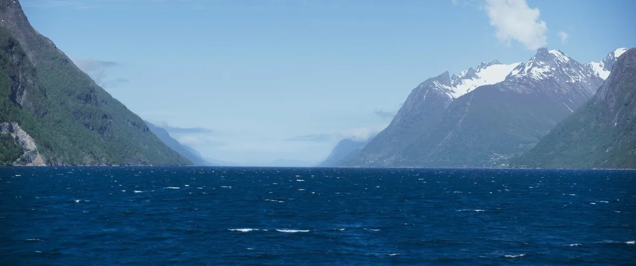 More Than Ever (2022) movie still: establishing — A wide view of a deep blue fjord flanked by steep green mountains. Snow-capped peaks rise on the…