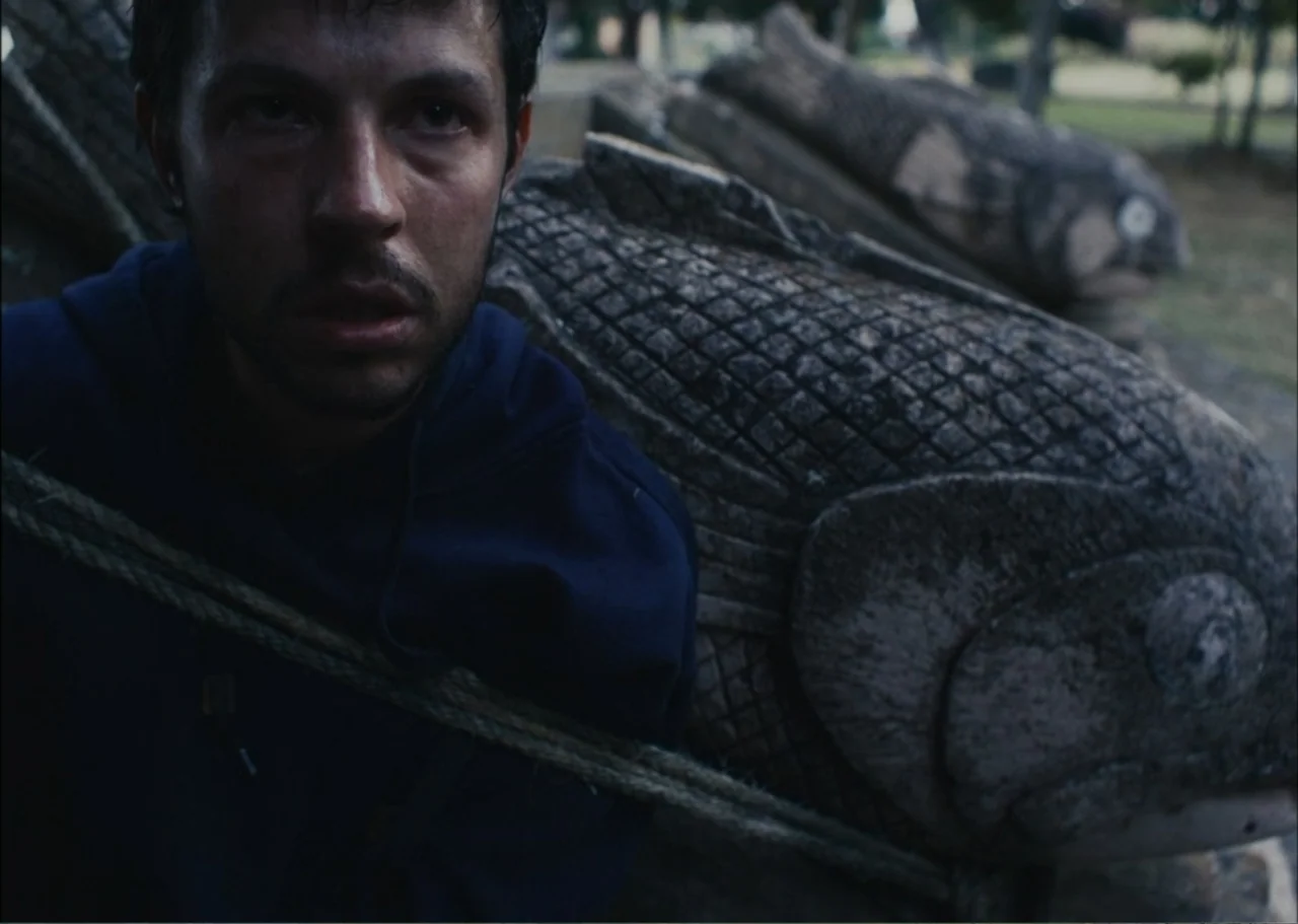 A Fisherman's Tale (1976) movie still: medium — A park setting features a stone sculpture of a fish near the man. A young man sits constrained by a…
