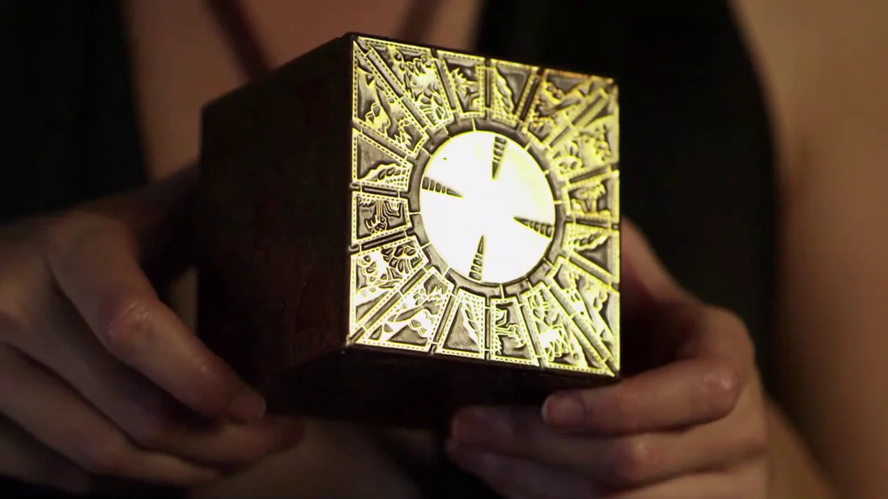 Hellraiser: Revelations movie still: closeup — An ornate dark metal cube with glowing engravings is held in two hands. The object radiates light…