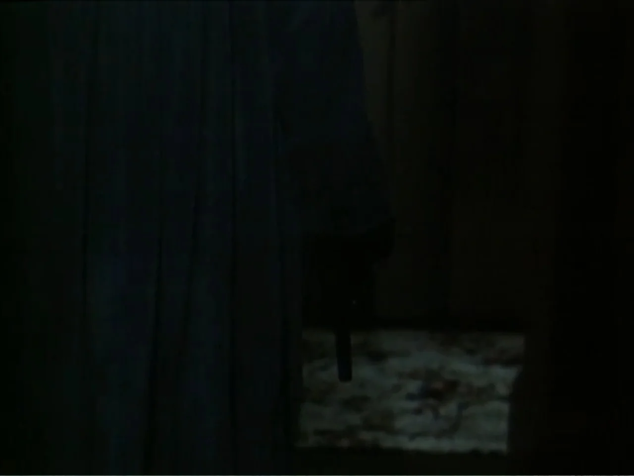Curtains (1995) movie still: medium — A dark, underexposed interior hallway features a figure wearing a floor-length dark fabric cloak. The…