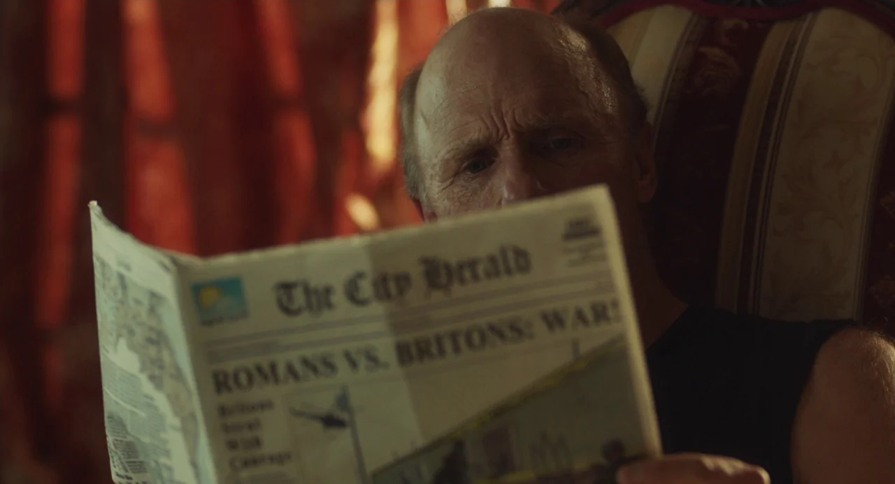 Cymbeline (2014) movie still: closeup — An elderly man reads a printed newspaper titled The City Herald. He sits in an upholstered chair inside a…