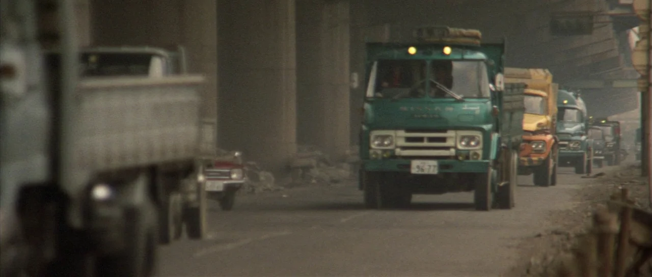 All Monsters Attack (1969) movie still: wide — A dusty asphalt road runs beneath a series of large concrete support pillars. Several heavy-duty…