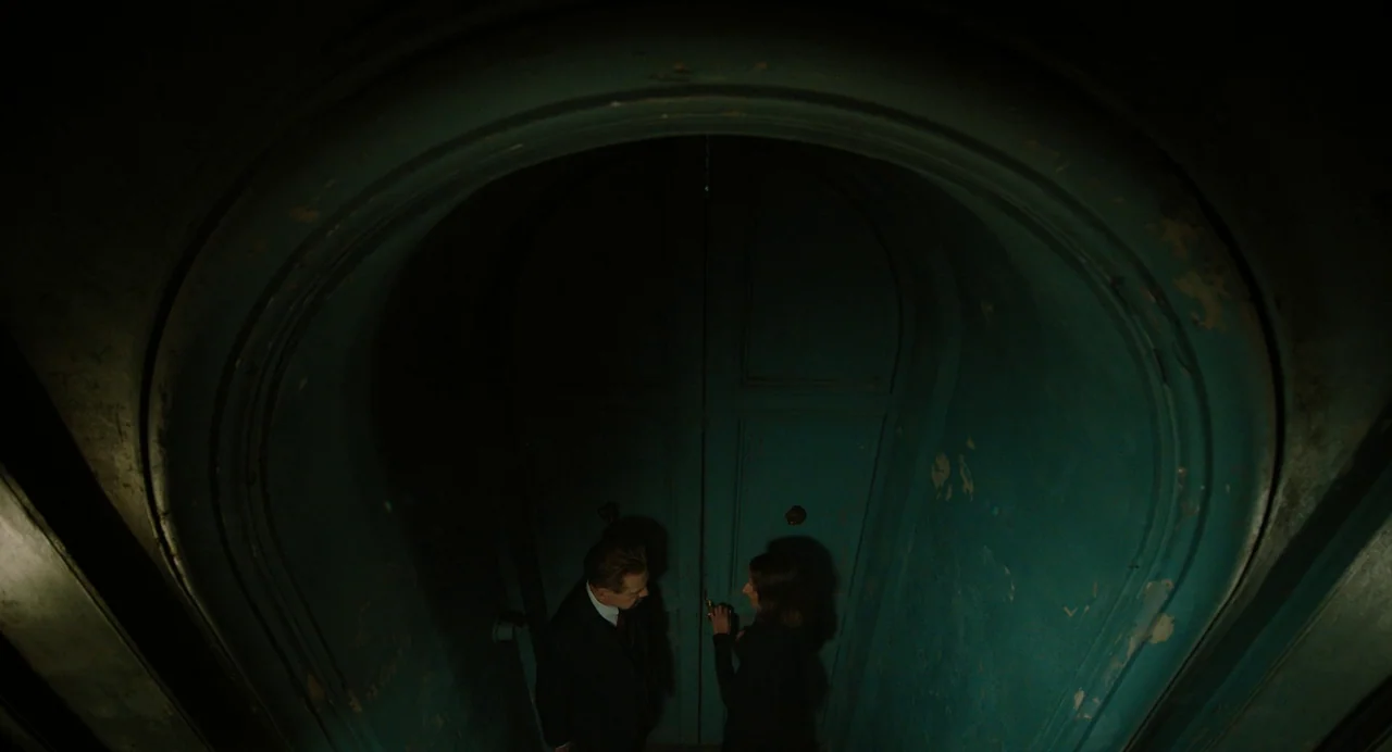 A Haunting In Venice (2023) movie still: wide — A man and a woman stand within a narrow, arched-ceiling vestibule with teal walls. They face a pair…