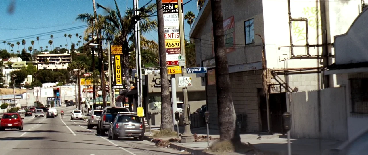 Little Birds (2011) movie still: wide — A Los Angeles commercial street features palm trees, utility poles, and mixed-use commercial buildings.…