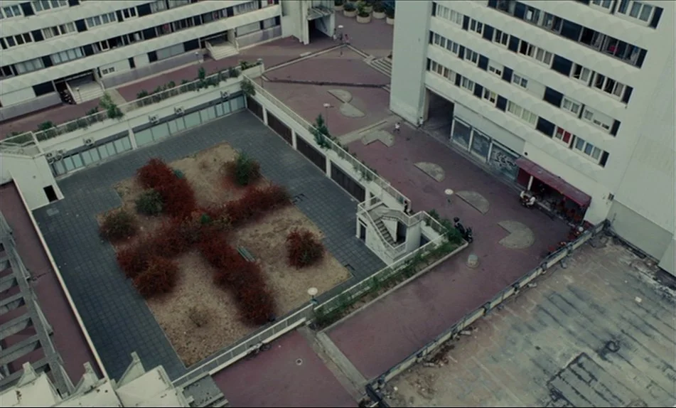 Hadewijch (2009) movie still: establishing — A high-angle view of a grey concrete apartment complex with an internal courtyard. Reddish-brown bushes…