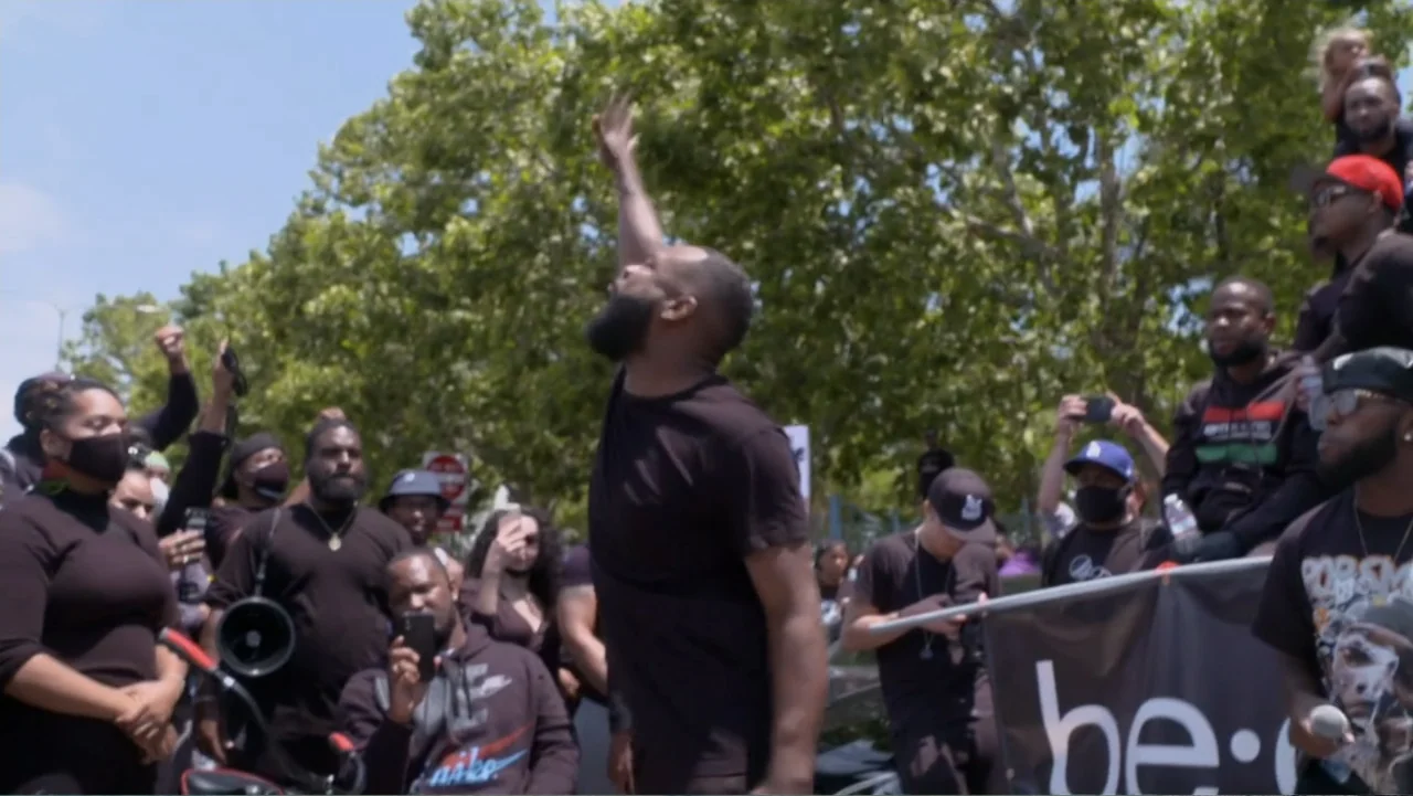 Between the World and Me (2020) movie still: medium — A group of people gather in a sunlit outdoor area during a public protest. A bearded man in a…