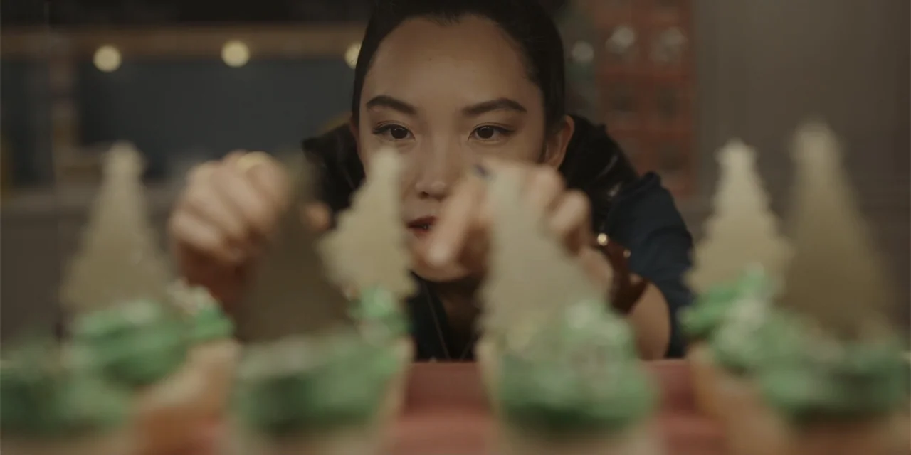 A Hollywood Christmas (2022) movie still: closeup — A bakery display counter holds several cupcakes decorated with green frosting and paper trees. A…