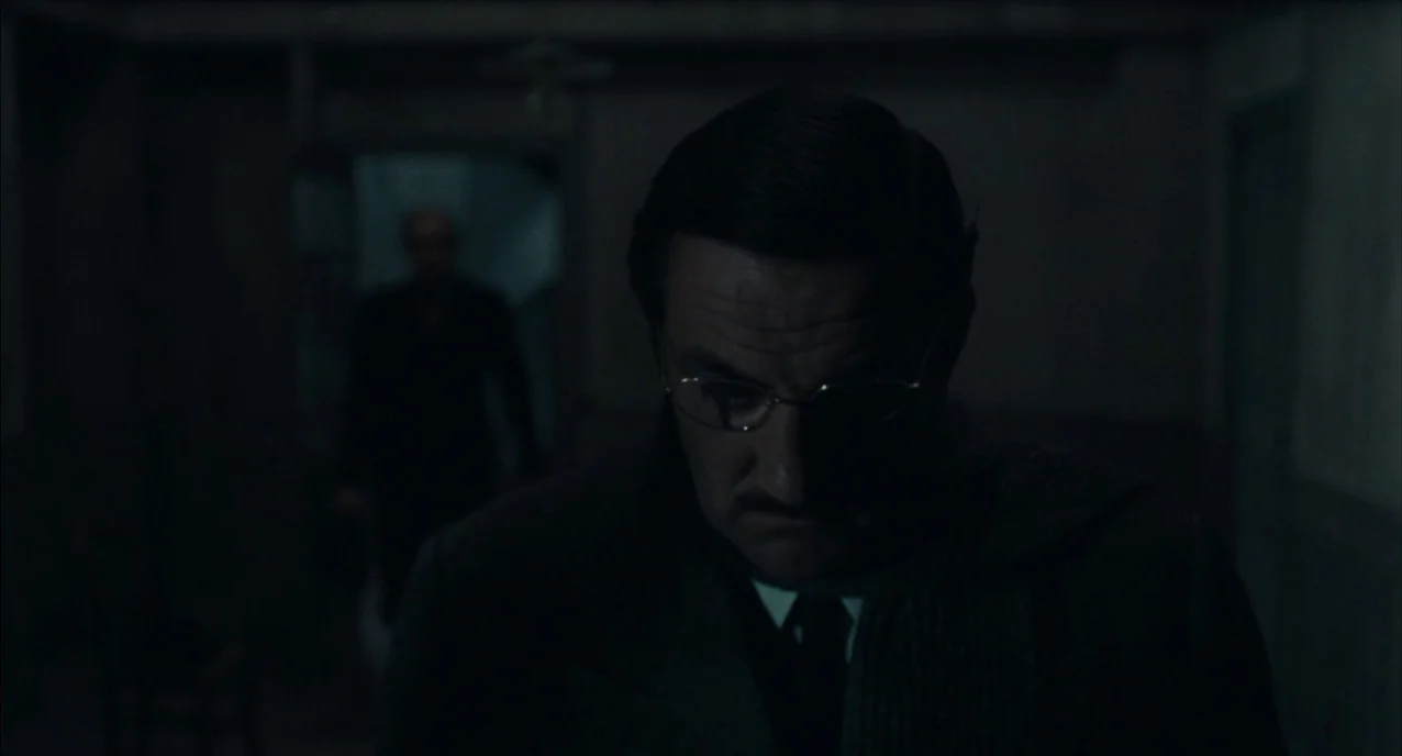 The Army of Shadows (1969) movie still: closeup — A dimly lit room with barely visible walls and a dark, indistinct background. A man stands in the…