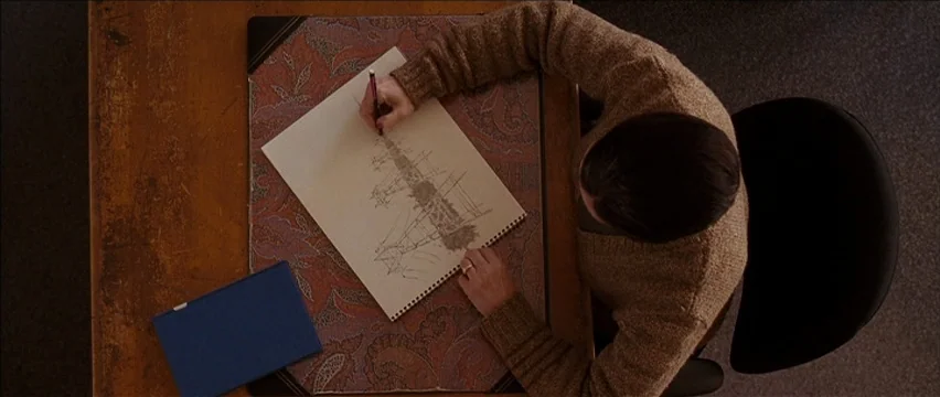 Snow Angels (2007) movie still: medium — A wooden desk features a floral patterned desk pad and a blue notebook. A man in a tan cardigan sketches a…