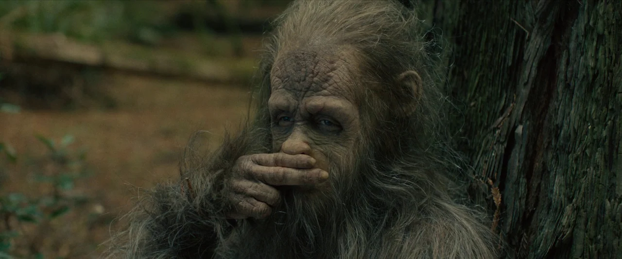 Sasquatch Sunset (2024) movie still: closeup — A hairy hominid creature sits at the base of a large, textured tree trunk. The subject rests a hand…