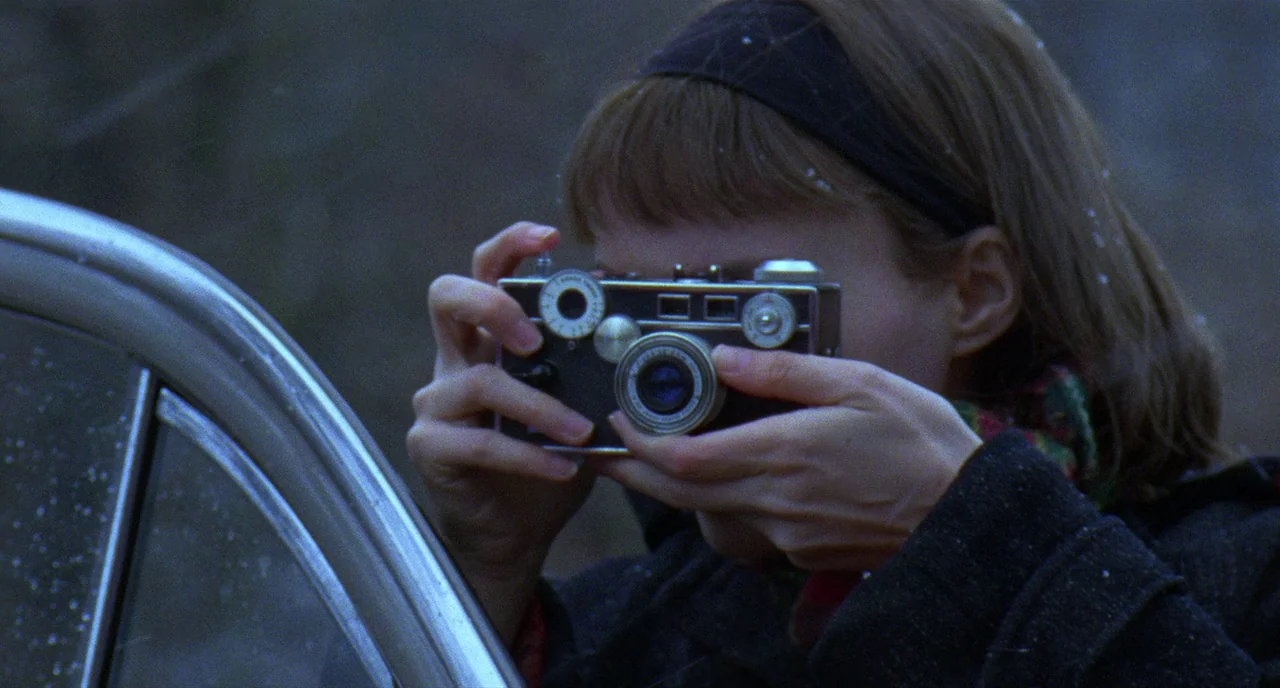 Carol (2015) movie still: closeup — A person sits inside a vehicle while aiming a vintage camera outward. Light snow falls around the dark exterior…