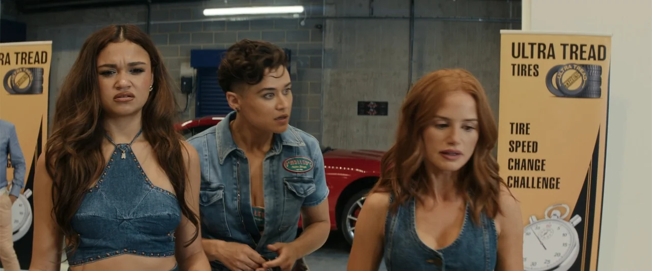Maintenance Required (2025) movie still: medium — Three young women stand inside a garage filled with automotive marketing materials. The group…