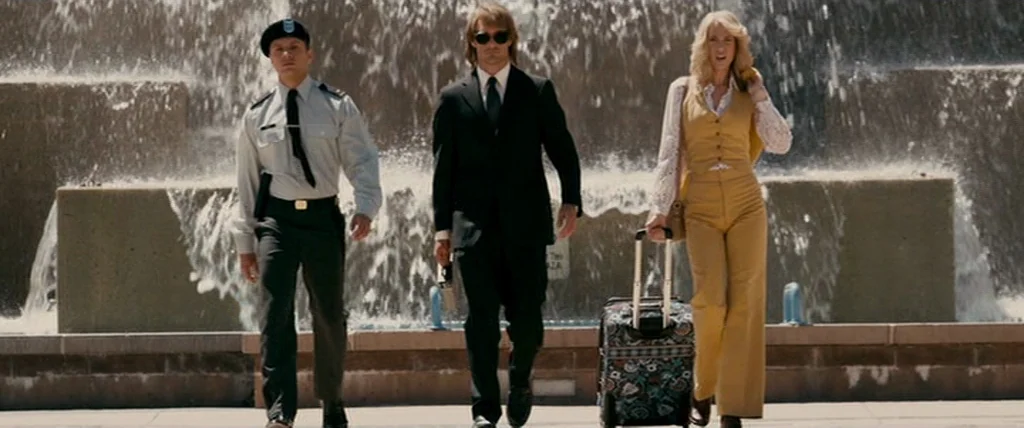 MacGruber (2010) movie still: fullbody — A concrete public square features a large water fountain spraying against a stone wall. Three people in…