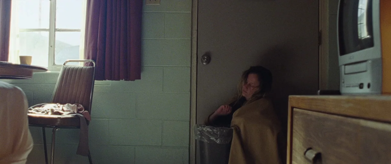 To Leslie (2022) movie still: wide — A spartan motel room features cinder block walls, a metal chair, and a small television. A woman sits on the…