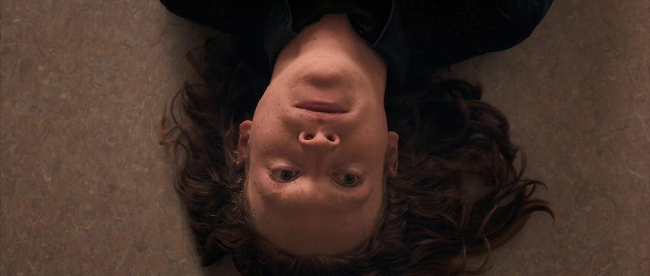 Byzantium (2012) movie still: closeup — A person lies flat on their back on a textured beige floor. They stare directly upward at the camera with a…