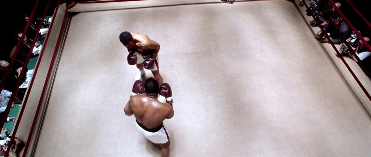 Ali (2001) movie still: wide — An empty, bright canvas boxing ring surrounded by red ropes and photographers. Two shirtless professional boxers…