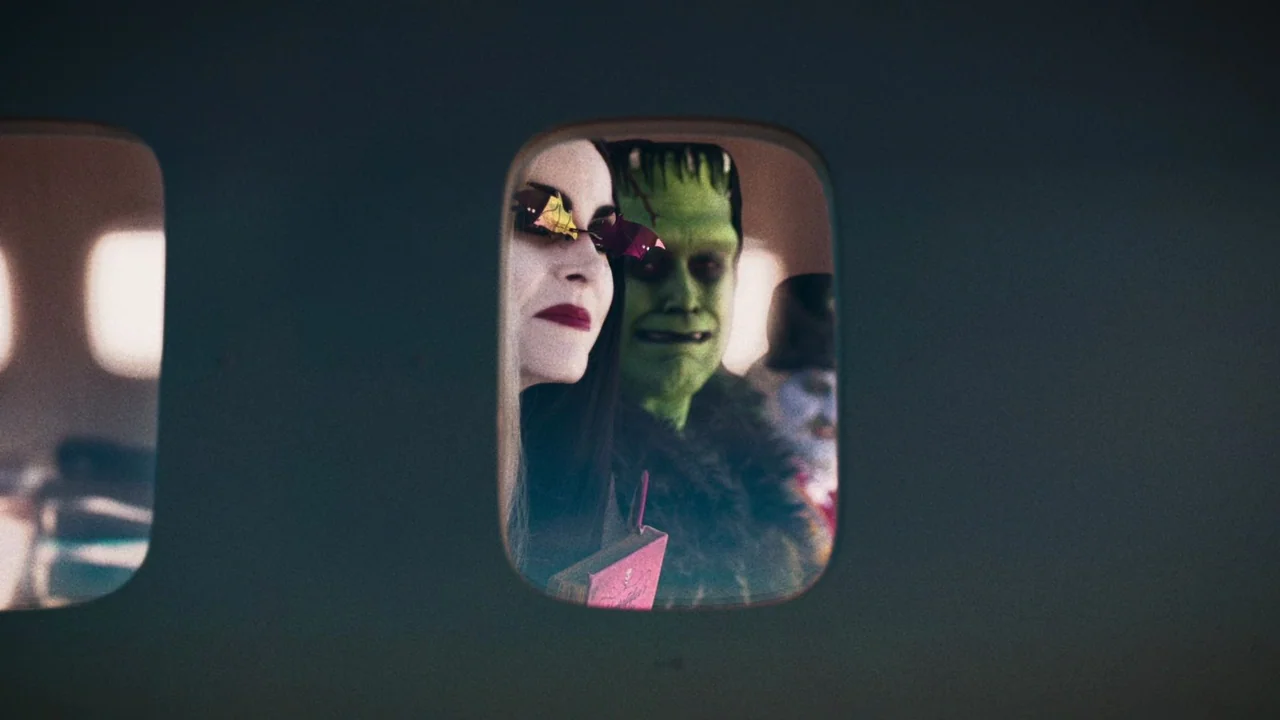 The Munsters (2022) movie still: medium — A view through an airplane window shows a pale woman and a green monster. They sit together in a row of…