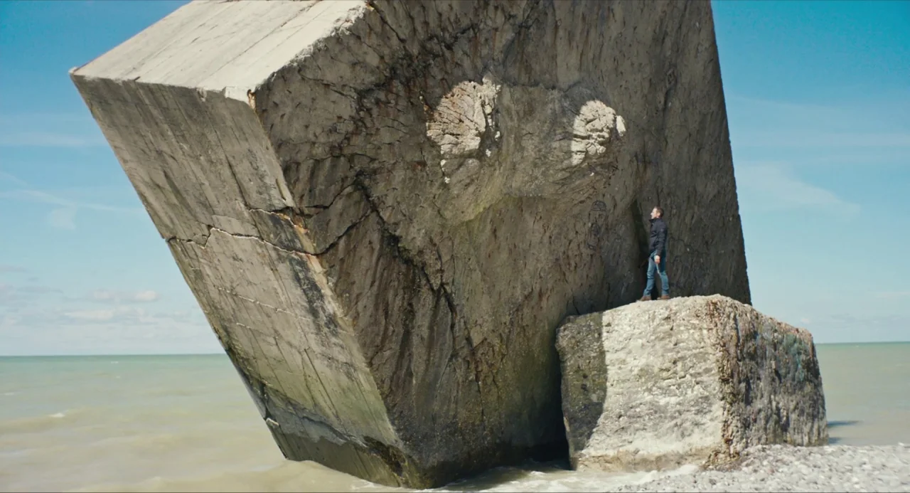 Submergence movie still: wide — A man stands atop a weathered concrete block on a pebbled beach. A massive tilted, reinforced concrete slab rises…