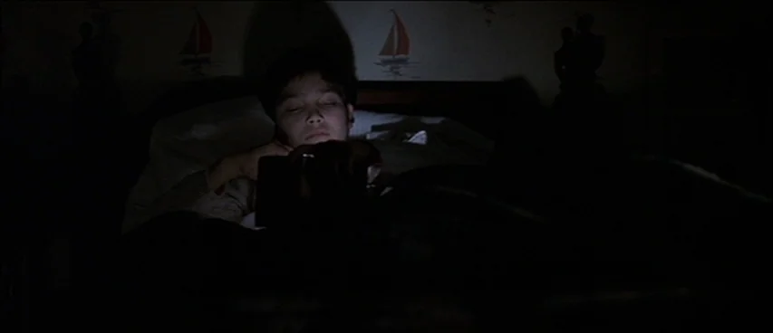 Road To Perdition (2002) movie still: medium — A dark bedroom with wall decor featuring small sailboat illustrations. A child lies in bed looking…