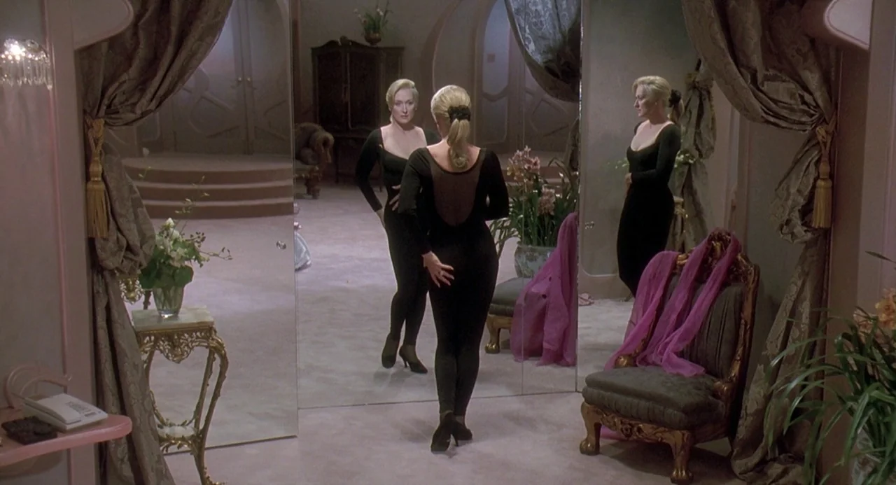 Death Becomes Her (1992) movie still: medium — A spacious room with pink carpet and ornate golden furniture reflects in large mirrors. A woman in a…