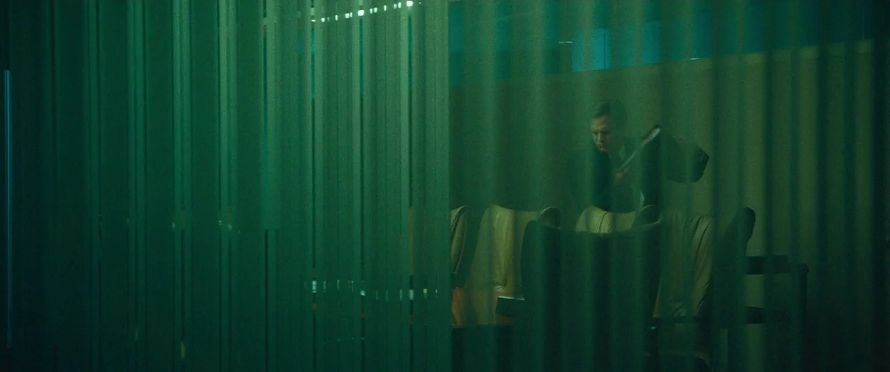 Dark Waters (2019) movie still: wide — A dark, green-toned boardroom viewed through vertical translucent blinds. A man in a suit stands alone near a…