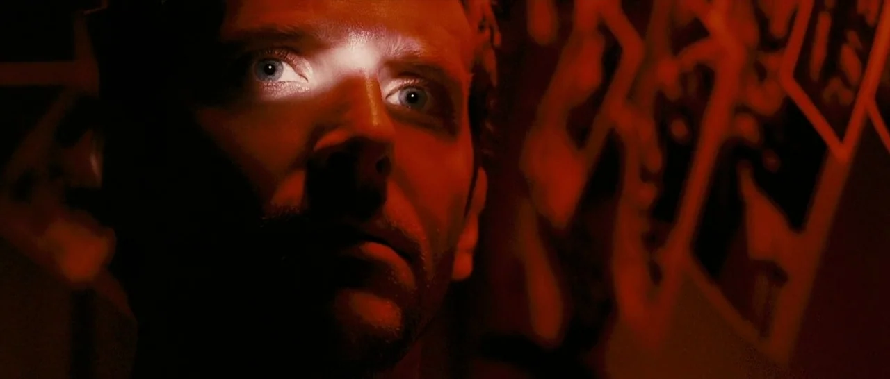 The Midnight Meat Train (2008) movie still: closeup — A dark indoor space with blurred, geometric patterns faintly visible on a wall. A man's face…
