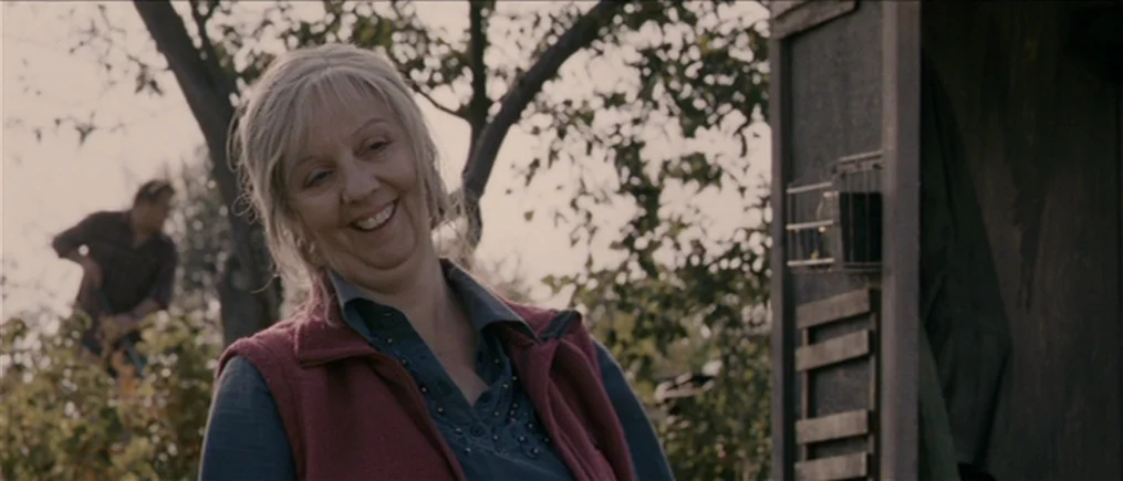 Another Year (2010) movie still: medium — An outdoor orchard with fruit trees and a wooden shed structure visible. A middle-aged woman smiles toward…
